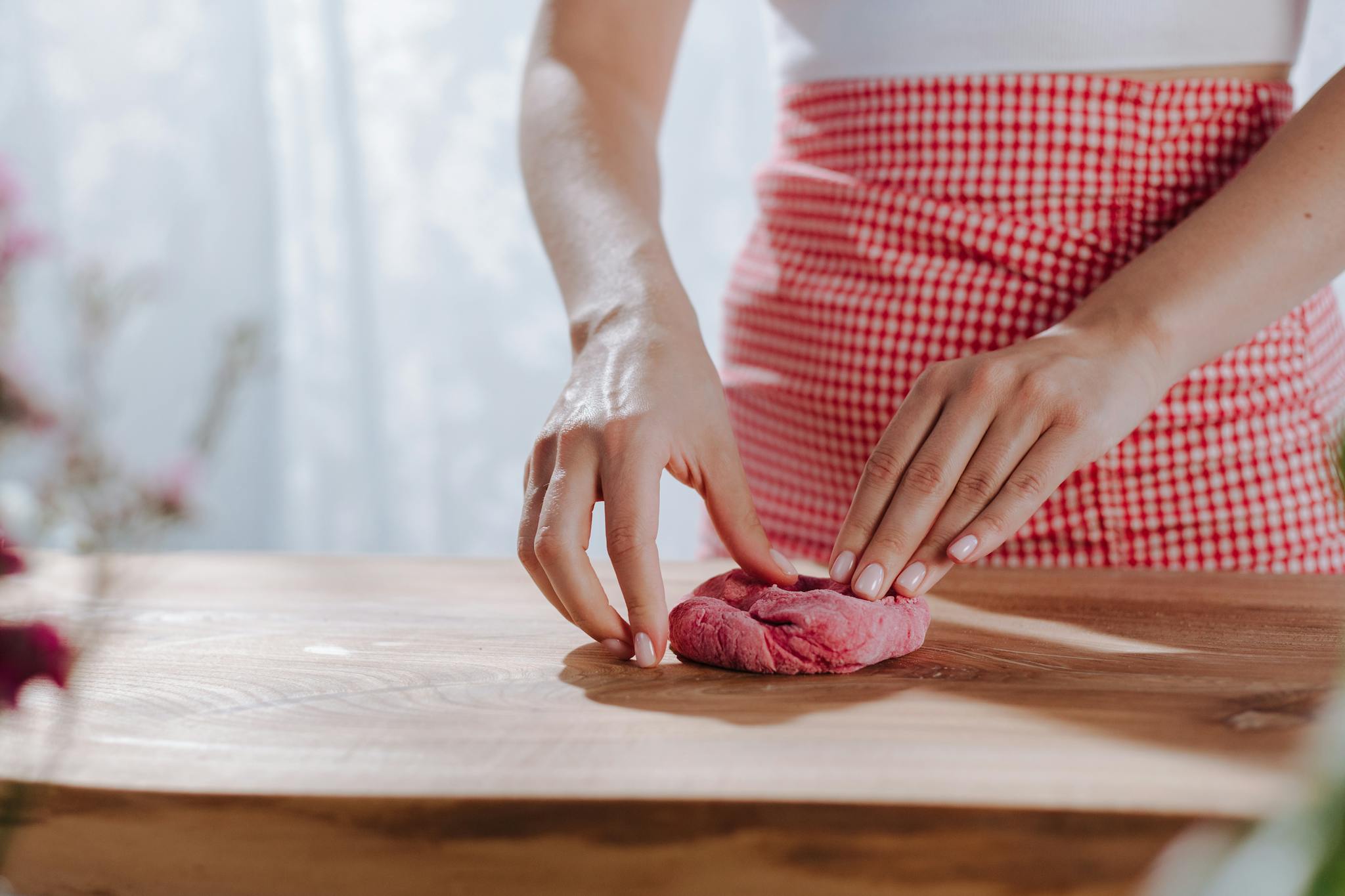 Woman in red gingham apron kneading vibrant red pasta dough on a wooden table.